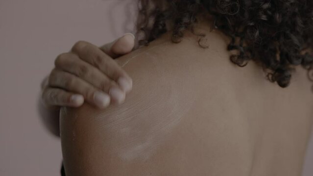 CU Back view, African-American female rubbing cream onto her shoulder. Studio shot, soft lighting. No make up, clean skin. Shot with ARRI Alexa Mini LF