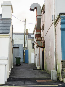 Narrow street or back alley with a restaurant air ventilation duct and one plastic wheelie bin. Muted white and grey color.