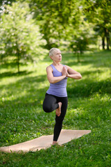 Middle-aged caucasian woman in sportswear practices yoga outdoors in a tree pose. Vrikshasana. The concept of aerial yoga, stretching, healthy lifestyle.Going in for sports in the city park.Side view.