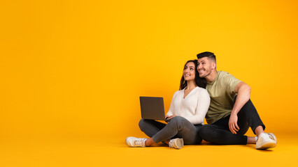 Happy amazed millennial arab man hugging lady with laptop, looking at empty space, sit on floor