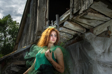 A girl in the form of a forest fairy near an old ruined wooden house.