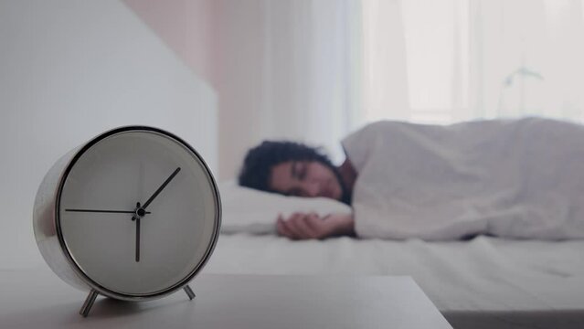 Close up shot of white alarm clock on table in bedroom with peaceful tranquil young woman sleeping on the side or shoulder comfortably on bed in blurred background. Daily early morning routine concept