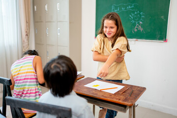 Caucasian girl stand in front of classroom and point to Asian boy look like angry with fun during learn in school.