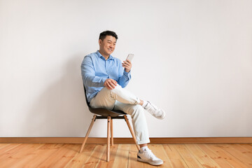 Excited middle aged asian man using smartphone, networking in social media, sitting on chair over white studio wall