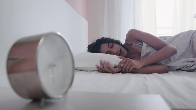 A young Indian Asian curly haired female or woman yawns and stretches her arms and wakes up or gets up from the cozy bed with a table or alarm clock in the foreground during morning hours