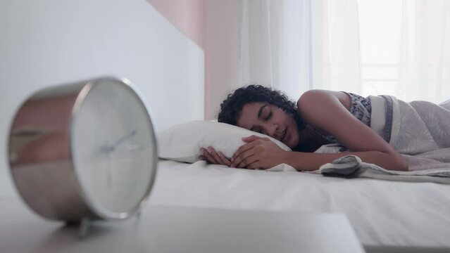 A young modern Indian Asian curly-haired female or woman yawns and stretches her hands while sleeping comfortably on a cozy bed or mattress with alarm or table clock in the foreground in the morning