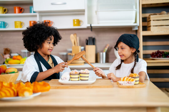 Two Little Girls, African American And Asian Girls Use Wooden Spoon And Fork To Play And Fun In Kitchen During Cooking Class.