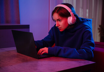 Teenager girl in white headphones playing video game on laptop while sitting at table indoors © Oksana Klymenko