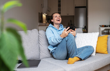 Woman using her mobile phone listening podcast and laughing while sitting on couch at home.