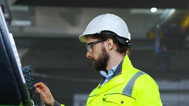 Close up of Caucasian workman in hardhat and yellow uniform pushing buttons on controlling point and running michine at plant. Indoors. Handsome man in goggles at controller computer in factory.