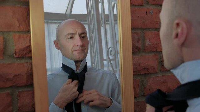 Male doing up his tie in a Windsor knot in mirror. A bald man getting ready for job interview or work. Happy and confident middle aged man.
