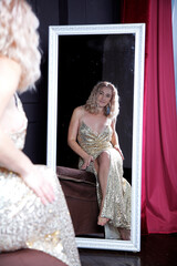 a woman in a golden dress looks in the mirror. A woman in a room in the bedroom in a beautiful festive dress, elegant. Blonde woman in the interior. Handsome man at home in krartir.