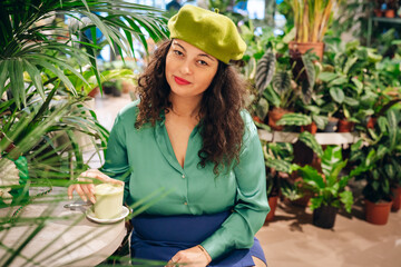 Stylish hipster woman drinking macha tea in green cafe. Trendy biophilic interior design with many green plants. Urban jungle - obrazy, fototapety, plakaty