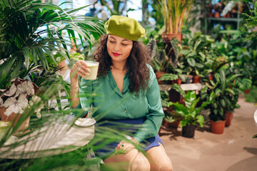 Woman in a green shirt drinking macha tea in cafe with many plants. Trendy biophilic interior design with many green plants. Urban jungle - obrazy, fototapety, plakaty