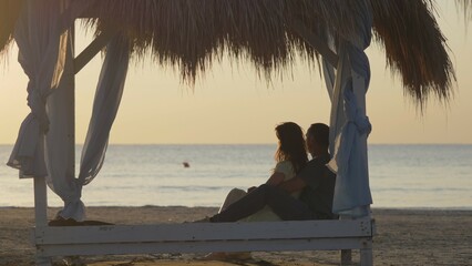 Couple of lovers sit on canopy, man and woman embraces admire serene sea © MEDIAIMAG