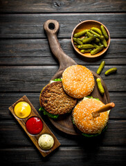 Burgers on a cutting Board with a knife and gherkins in a bowl.