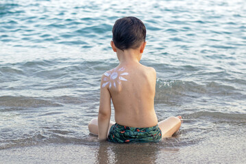 kid boy preschooler with drawn sun shape from cream lotion on shoulder against sea ocean tan red...