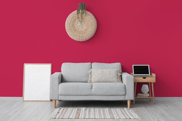 Comfortable sofa, blank photo frame and table with modern laptop near viva magenta wall in room