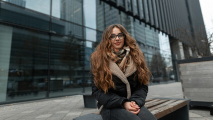 Happy beautiful fashionable woman with glasses in stylish fashion winter clothes with a scarf, jacket and jeans sits and freezes on a bench in the modern city