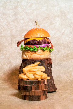 Hamburguesa artesanal con queso americano, tocineta y vegetales sobre troncos de madera y papas fritas en el centro sobre troncos con un fondo c&aacute;lido.