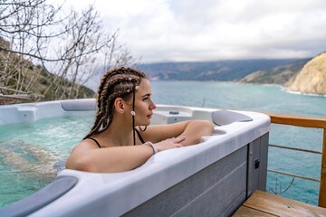 Outdoor jacuzzi with mountain and sea views. A woman in a black swimsuit is relaxing in the hotel pool, admiring the view - obrazy, fototapety, plakaty