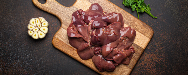 Raw chicken liver on wooden cutting board top view on dark rustic concrete background kitchen table with parsley and garlic. Healthy food ingredient, source of iron, folate, vitamins and minerals