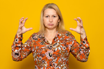 Frustrated blonde woman in print dress with furious expression, raising claws in aggression, experiencing strong emotions. Indoor studio shot isolated on orange background