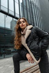 Beautiful fashionable woman model with a curly hairstyle in fashion winter clothes with a down jacket and a knitted sweater with jeans posing near a business building in the city