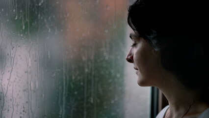 Woman standing by window during rainy day looking outside watching rain