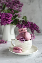 Vertical spring composition. White coffee cup with macaroons and a bouquet of lilacs on a light gray background. Festive concept or concept for a good morning wish. Selective focus. 