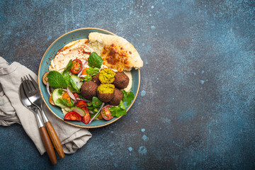 Middle Eastern Arab meal with fried falafel, hummus, vegetables salad with fresh green cilantro and mint leaves, pita bread in ceramic bowl on stone rustic background top view, space for text