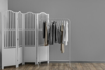 Rack with stylish female clothes and folding screen near grey wall