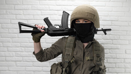 a girl soldier in a helmet and military ammunition with a Kalashnikov assault rifle against a brick wall