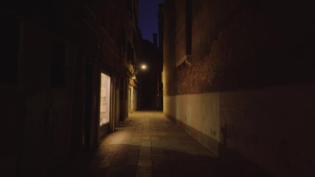 Long narrow dark historical street between old buildings illuminated by single bright shop window and lamp as popular attraction in Venice Italy