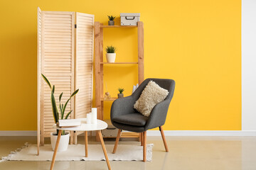 Interior of living room with folding screen, armchair and table near yellow wall