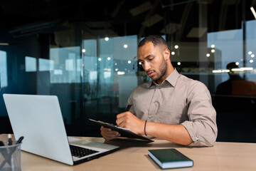 Serious concentrated businessman writing document while sitting in office using laptop for online...
