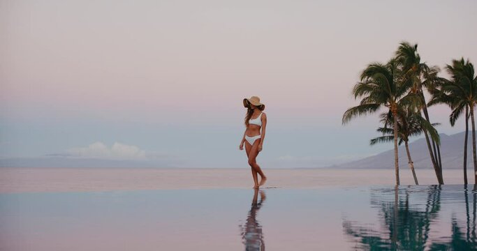 Beautiful woman walking along the edge of luxury infinity pool at tropical resort spa at sunrise