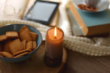 Vintage books, reading glasses, e-reader, cup of tea, bowl of biscuits and lit candle on the table. Selective focus.