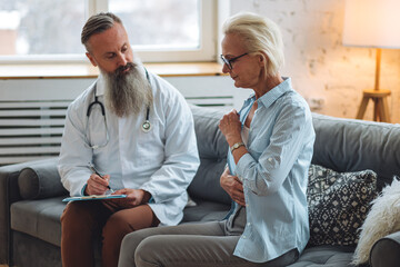Obraz premium Male doctor visiting attractive senior woman at home for consultation, medical service and support, geriatric help, prescribing illness treatment. Clinic or hospital appointment, cancer recovery