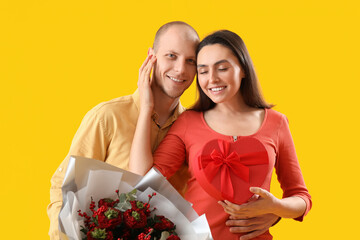 Young couple with bouquet of flowers and gift on yellow background. Valentine's Day celebration