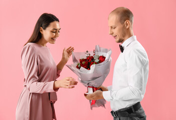 Young man with flowers and ring proposing to his girlfriend on pink background. Valentine's Day celebration