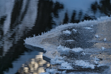 Frozen and icy part of the river in winter makes a layer of ice near the shore, snowflakes are like fluffy needles, reflection of the winter forest in the water of the river