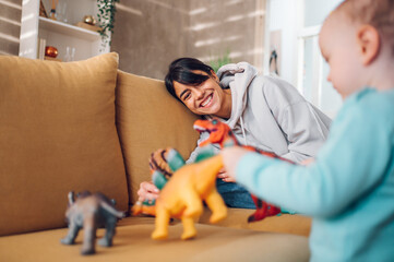 Mother and toddler son playing with dinosaurs toys at home on the couch