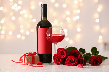 Glass of wine, rose flowers, bottle and gift on white table against blurred lights. Valentine's Day...
