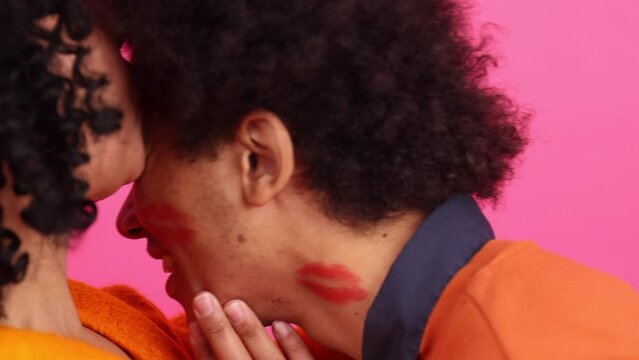 Portrait of African American, young boyfriend and girlfriend on pink isolated background. Close-up view of a woman leaving lipstick marks on a face of her man. High quality 4k footage