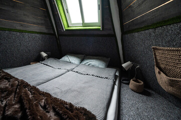 Double bed on carpet floor in bedroom of wooden cabin. Pyramid shape bungalow from inside. Small room, angled walls, window light, made bed. Handmade interior of wooden cabin.
