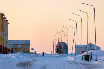 People walk along the snow-covered street of the northern settlement in the Arctic. Buildings,...