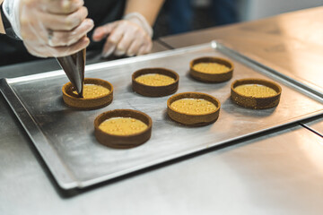 Unrecognisable baker piping chocolate ganache into filled mini tart crusts. Professional baking process. Assembling dessert. Horizontal indoor close-up shot. High quality photo