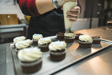 Female baked in black apron piping white frosting on chocolate cupcakes on siler tray. Professional baking. Horizontal indoor shot. High quality photo