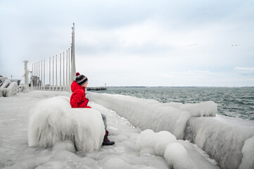 Child in red coat sitting on icy bench on frozen pier in winter.